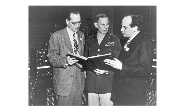 Black and white photo of John Mauchly And J Presper Eckert looking at an open book with a person in military uniform