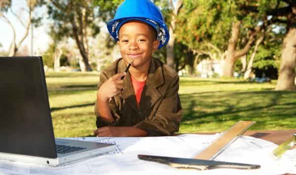 Image of a kid wearing a hardhat and holding a pencil at an outdoor table with a laptop, T-square and blueprints. 