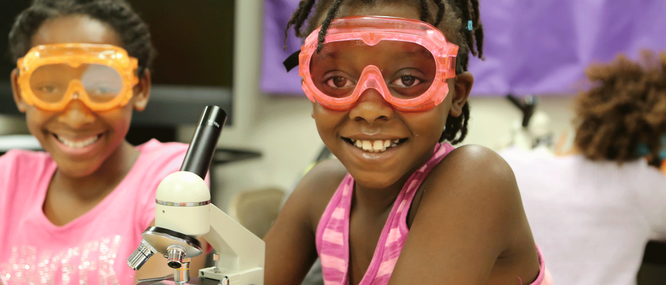 Photograph of two girls in goggles and lab equipment looking through a microscope