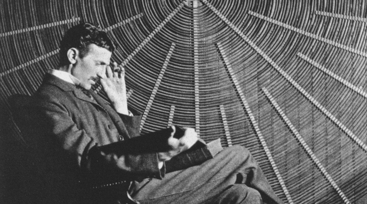 Black and white photograph of Nikola Tesla in front of the spiral coil of his high-voltage Tesla coil transformer