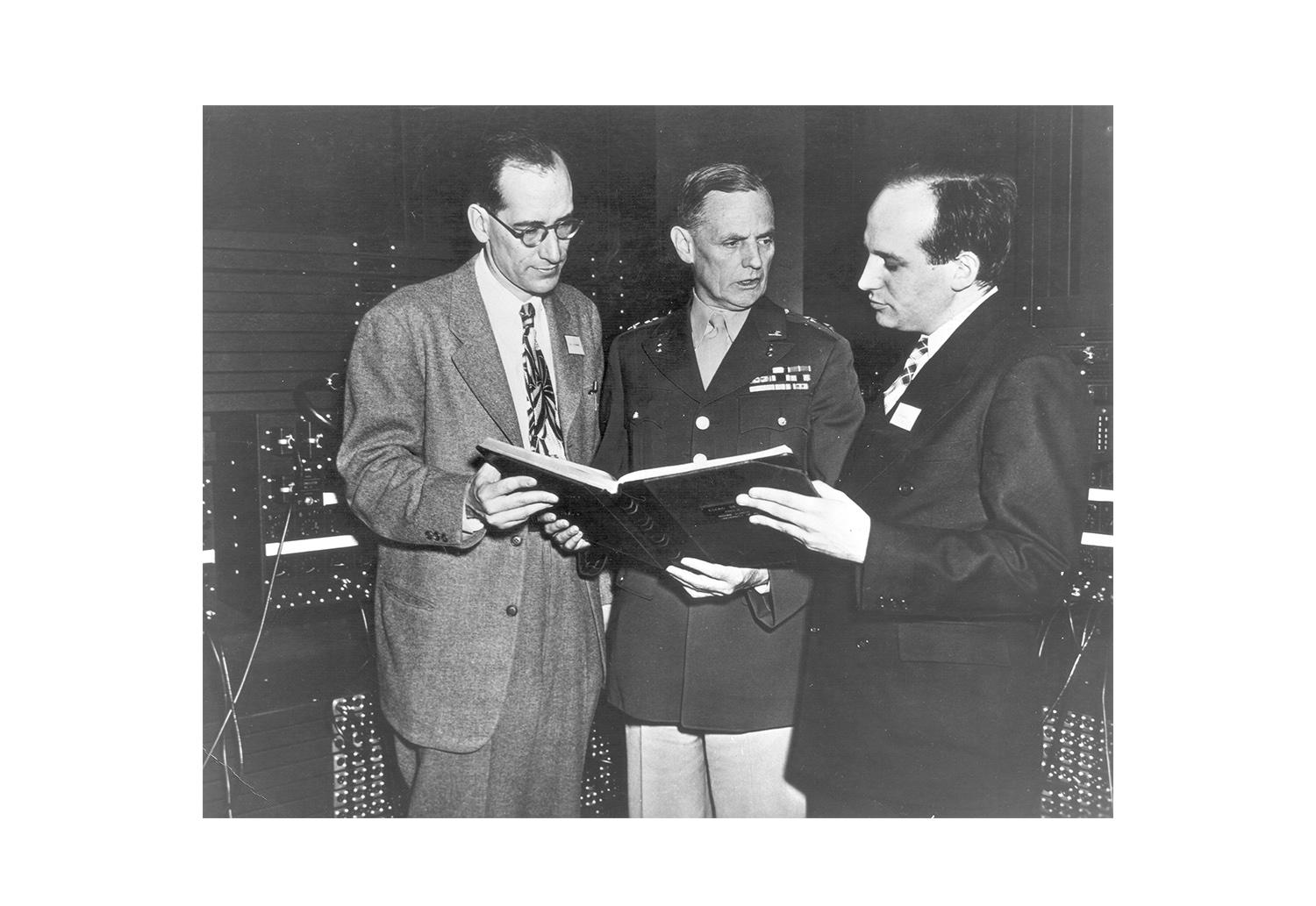 Black and white photo of John Mauchly And J Presper Eckert looking at an open book with a person in military uniform